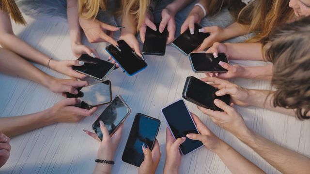 63354096_people-holding-their-smartphones-around-a-white-table-using-their-devices-and-showing-a-sense-of-community